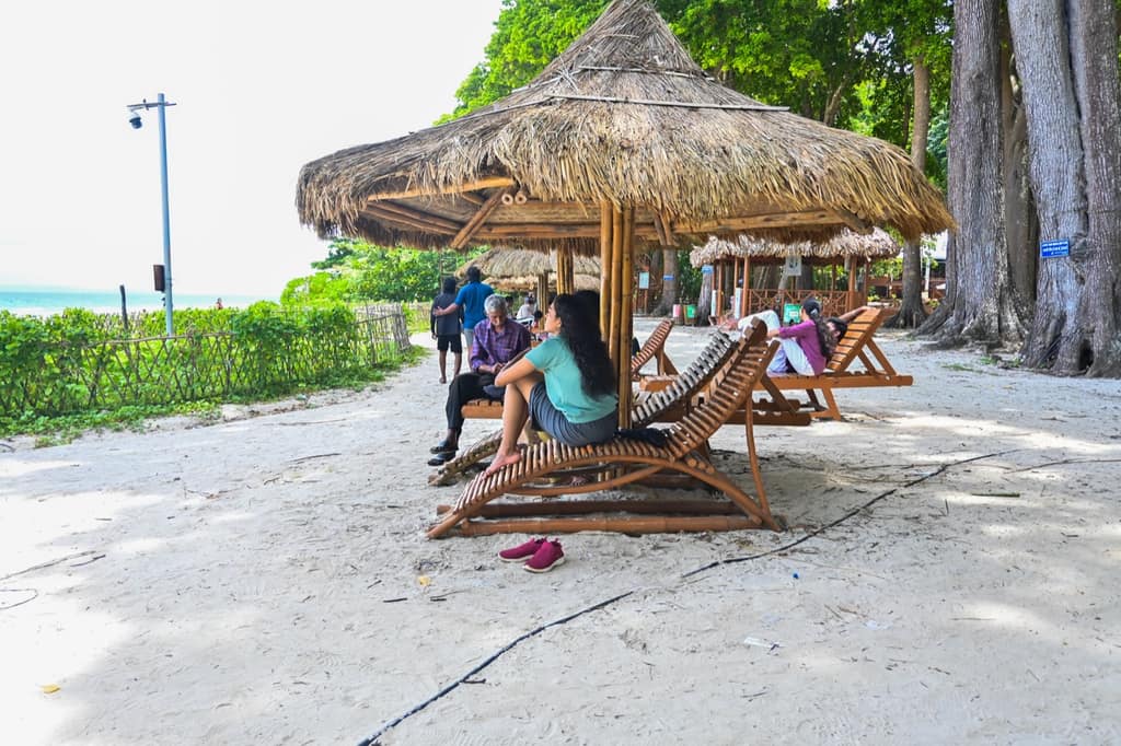Shaded wooden loungers under thatch umbrellas at the treeline of Radhanagar Beach